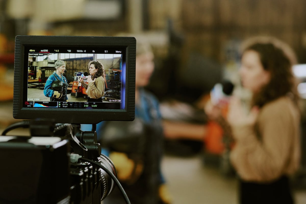Two women being filmed by a professional camera crew in an industrial workshop while a monitor displays their conversation
